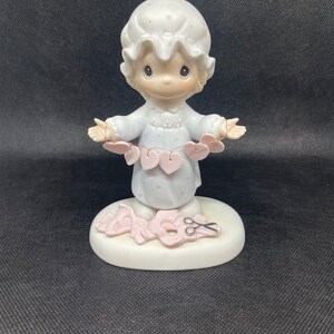 May include: A porcelain figurine of a girl in a nightgown holding a string of pink hearts. She is standing on a base with a pair of scissors and a heart cutout.