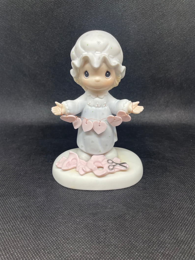 May include: A porcelain figurine of a girl in a nightgown holding a string of pink hearts. She is standing on a base with a pair of scissors and a heart cutout.