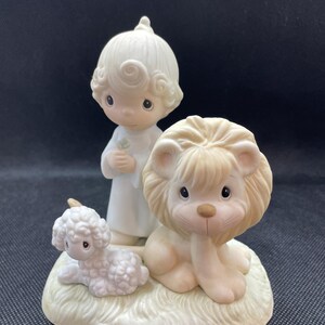May include: A ceramic figurine featuring a child, a lion, and a lamb. The child is dressed in white and holds a small object. The lion and lamb are light beige and white, respectively, and rest on a grassy base.