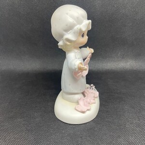 May include: A porcelain figurine of a young girl wearing a white nightgown and a white bonnet. She is holding a pink heart in her hand and is surrounded by pink hearts on the base. The figurine is part of the Precious Moments collection.