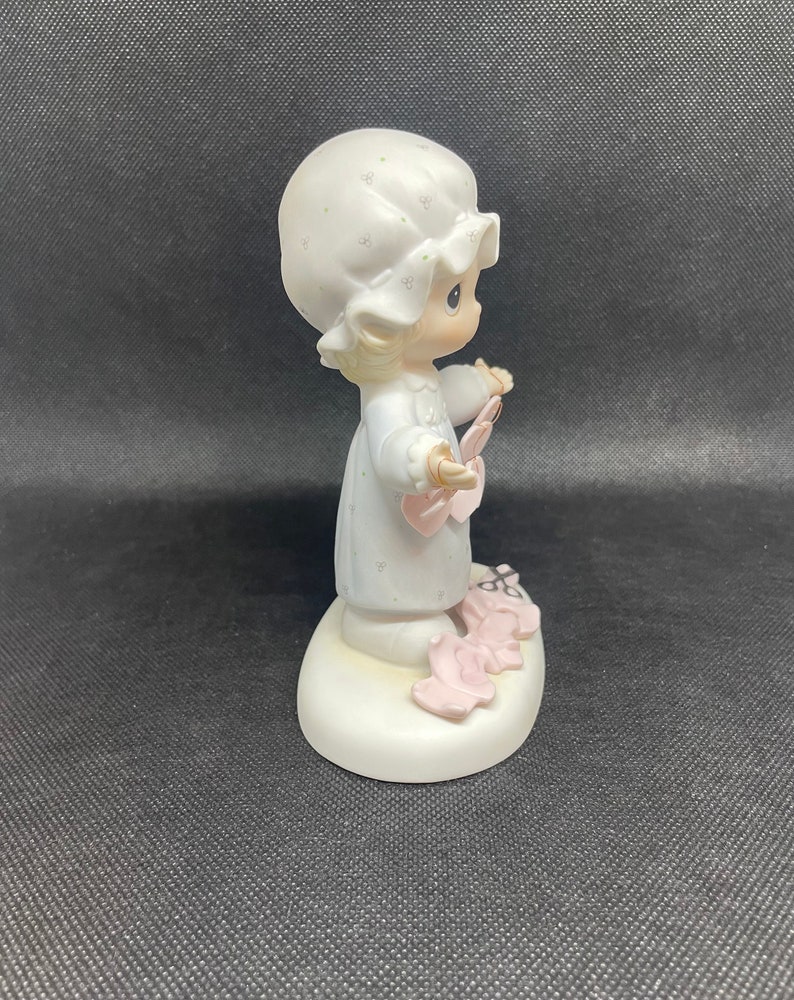 May include: A porcelain figurine of a young girl wearing a white nightgown and a white bonnet. She is holding a pink heart in her hand and is surrounded by pink hearts on the base. The figurine is part of the Precious Moments collection.