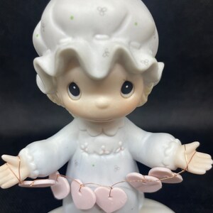 May include: A porcelain figurine of a girl wearing a white nightgown and a white bonnet. She is holding a string of pink hearts. The figurine is standing on a base with pink hearts.