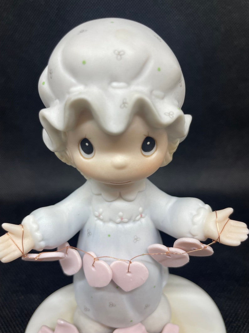 May include: A porcelain figurine of a girl wearing a white nightgown and a white bonnet. She is holding a string of pink hearts. The figurine is standing on a base with pink hearts.
