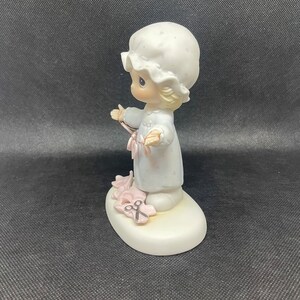 May include: A porcelain figurine of a little girl in a white nightgown and a white bonnet holding a pair of pink scissors. The figurine is standing on a white base with pink flowers and a pair of scissors.