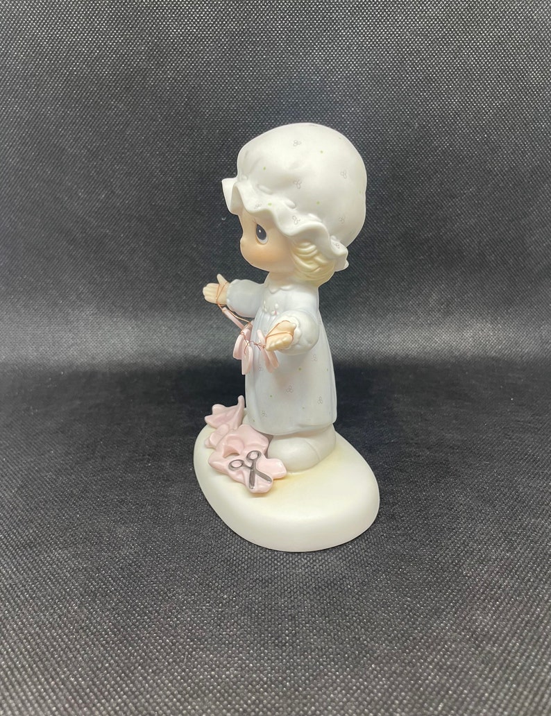 May include: A porcelain figurine of a little girl in a white nightgown and a white bonnet holding a pair of pink scissors. The figurine is standing on a white base with pink flowers and a pair of scissors.