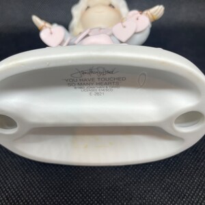 May include: A white porcelain figurine of an angel with blonde hair and a pink and blue dress. The angel is holding out its arms with hearts in its hands. The base of the figurine has the text "You have touched so many hearts" and "Jonathan & David 1983, Licensee Enesco, E-2821".