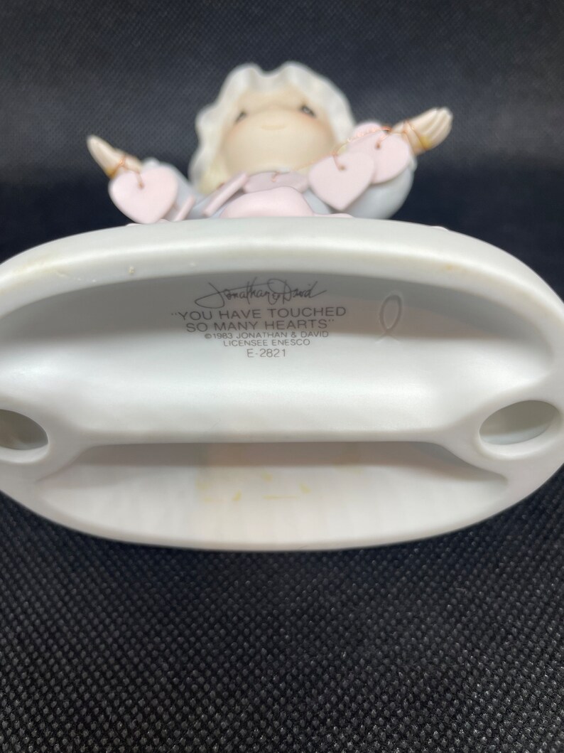 May include: A white porcelain figurine of an angel with blonde hair and a pink and blue dress. The angel is holding out its arms with hearts in its hands. The base of the figurine has the text "You have touched so many hearts" and "Jonathan & David 1983, Licensee Enesco, E-2821".