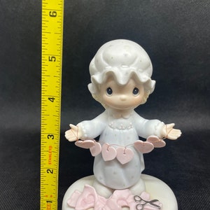 May include: A porcelain figurine of a girl in a nightgown holding a string of pink hearts. She is standing on a base with a pair of scissors and a pink ribbon.