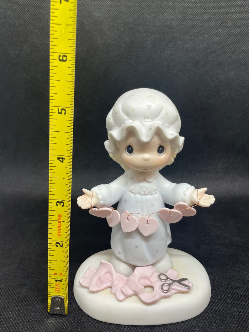 May include: A porcelain figurine of a girl in a nightgown holding a string of pink hearts. She is standing on a base with a pair of scissors and a pink ribbon.