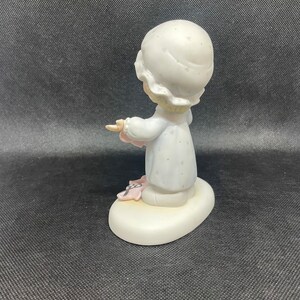 May include: A porcelain figurine of a girl wearing a white nightgown with a floral pattern and a white nightcap. She is holding a pink blanket and has a sweet expression on her face. The figurine is standing on a white oval base.