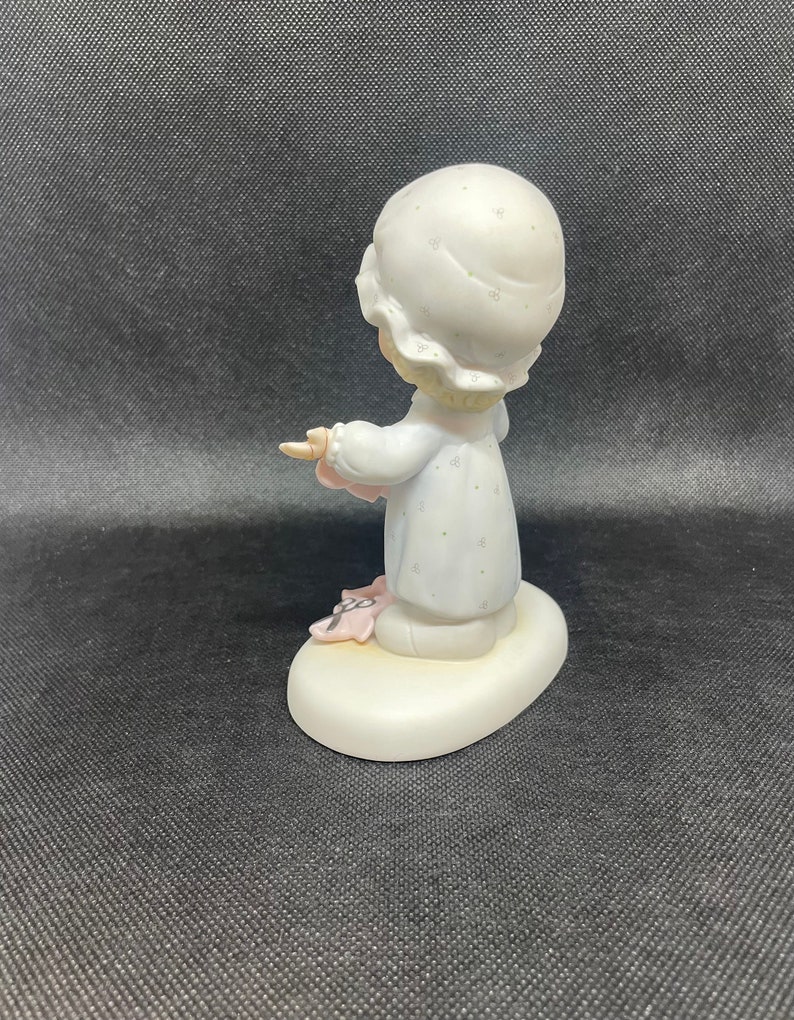 May include: A porcelain figurine of a girl wearing a white nightgown with a floral pattern and a white nightcap. She is holding a pink blanket and has a sweet expression on her face. The figurine is standing on a white oval base.
