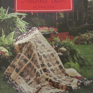 May include: A quilt with a beige background and a pattern of squares with dark brown and black images. The quilt is draped over a wrought iron chair in a garden setting. The text "Azalea Trail" is displayed on a red sign.