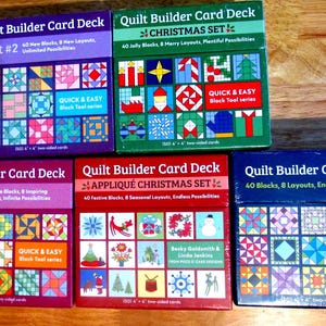 May include: A selection of Quilt Builder Card Decks, showcasing different sets with vibrant quilt block patterns. The boxes are in purple, green, red, and blue, with text detailing the number of blocks and layouts. Each box includes the "Quick & Easy Black Tool" series.