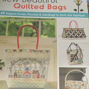 May include: The cover of a book, "Sew Beautiful Quilted Bags", displays a light blue quilted tote bag with red handles. The bag has a stitched design of houses and trees. Other bags are shown with various designs. The book is by Akemi Shibata.