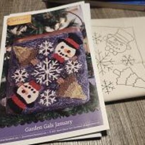 Peut inclure: Un modèle d'artisanat intitulé Garden Gals January présentant un motif carré violet avec deux bonhommes de neige portant des chapeaux noirs, deux sapins marron et des flocons blancs. À côté se trouve un tissu blanc avec un dessin au trait noir.