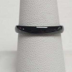 May include: A close-up of a black ring. The ring is a solid, glossy black color and appears to be made of a smooth material. The ring is displayed on a white surface.
