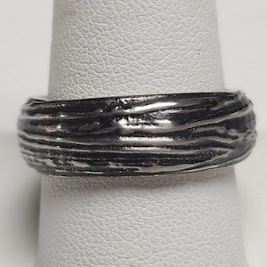 May include: A silver-colored ring with a textured, wood-grain-like design. The ring is a band style, with a slightly rounded profile. The surface has horizontal ridges, creating a unique, organic appearance. The ring is displayed on a white background.