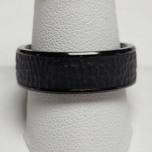 May include: A black ring with a hammered texture band. The ring is shown on a white display stand. The band has a textured, hammered appearance, and the edges are smooth and shiny.