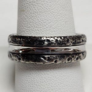 May include: A silver ring with a textured, dark gray outer band and a smooth, shiny silver inner band. The ring is displayed on a white surface.