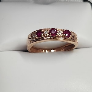 May include: Rose gold ring featuring four round, deep red rubies and three small diamonds. The band has an intricate floral design. The ring is set against a white background.