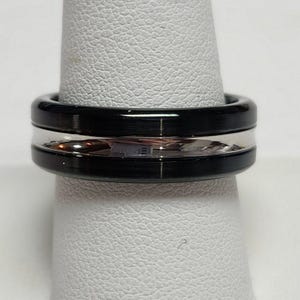 May include: A black and silver ring. The ring has a wide, black band on the top and bottom, with a silver band in the middle. The ring is displayed on a white surface.