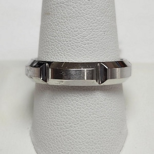 May include: A silver-colored ring with a geometric design. The ring has a flat top and beveled edges, with two rectangular cutouts. The ring is displayed on a white ring holder.