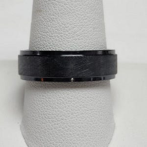 May include: A black ring with a brushed metal finish. The ring is shown on a white surface. The ring has a flat, wide band with a textured surface and a polished edge.