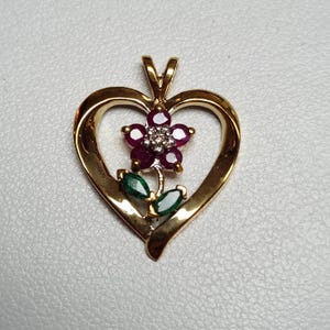 May include: A gold heart-shaped pendant featuring a central flower motif. The flower has five deep red ruby petals surrounding a small cluster of clear diamonds. Two marquise-cut green emerald leaves are positioned below the flower petals.