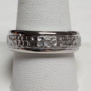 May include: A silver ring with a hammered texture band. The ring is shown on a white background. The ring has a polished, smooth edge on the top and bottom of the band.