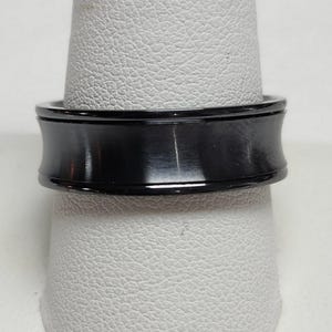 May include: A black ring with a concave center and polished edges. The ring is displayed on a white surface, showcasing its sleek, modern design. The ring's dark color contrasts with the light background.