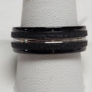 May include: A black ring with a hammered texture and a silver band. The ring is on a white surface.