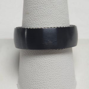 May include: A black ring with a brushed metal finish. The ring has a smooth, rounded band and a textured inner edge. The ring is displayed on a white ring holder.