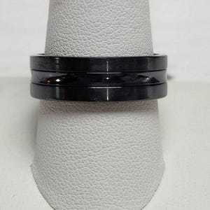 May include: A black ring with a grooved center band. The ring is made of a dark, shiny material and is displayed on a white surface. The ring has a modern, minimalist design.