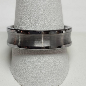 May include: A silver-colored ring with a brushed metal center band and polished edges. The ring has a unique, curved design and is displayed on a white surface. This piece of jewelry is suitable for everyday wear.