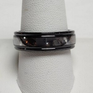 May include: A black and silver ring with a polished finish. The ring has a black outer band and a silver inner band. The ring is displayed on a white ring holder. The ring is a jewelry item.
