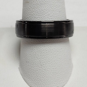 May include: A black ring with a brushed metal finish. The ring is shown on a white display stand. The ring has a flat top and a smooth, polished band. The ring is a simple, modern design.