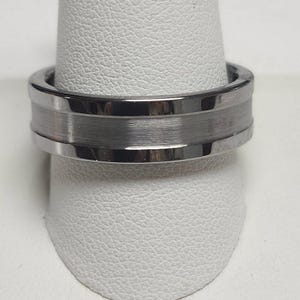 May include: A silver-colored ring with a brushed metal center band. The ring is displayed on a white stand, showcasing its polished edges and textured design. This piece of jewelry is suitable for everyday wear.