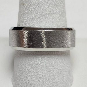 May include: A silver-colored ring with a brushed metal finish. The ring has a flat top and a polished, shiny band. The ring is displayed on a white surface.