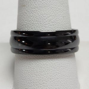 May include: A black ring with a polished, glossy finish. The ring has a wide band with a grooved design, creating a layered appearance. The ring is displayed on a white surface.