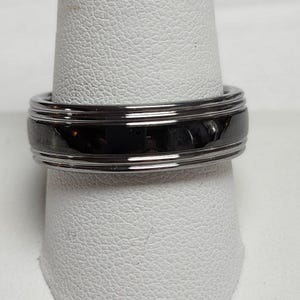 May include: A black and silver ring with a polished finish. The ring has a wide band with a black center and silver edges. The ring is displayed on a white surface.