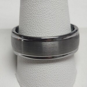 May include: A close-up of a silver-colored ring with a brushed, gray center band. The ring is displayed on a white, textured surface. The ring has a polished, shiny finish on the outer edges.