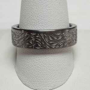 May include: A dark gray ring with an engraved floral pattern. The ring is made of a solid metal and has a wide band. The engraved design covers the entire outer surface of the ring.
