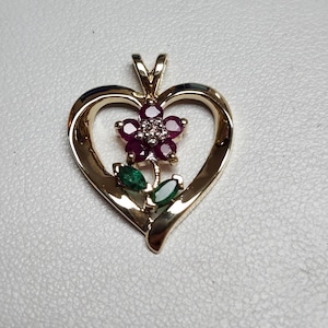 May include: A gold-colored pendant in the shape of a heart. The pendant features a flower with ruby petals and a diamond center, along with two emerald-colored leaves. The pendant is designed to be worn as jewelry.
