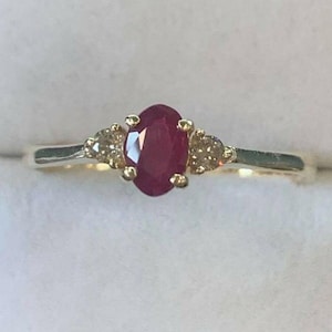 May include: A gold ring featuring an oval, deep red ruby center stone, flanked by two smaller, round, light brown gemstones. The ring's band is a thin, polished gold.