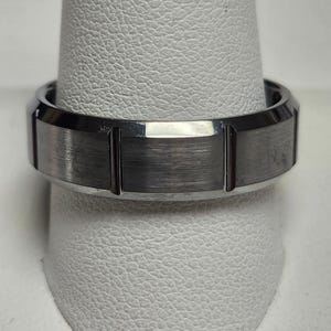 May include: A silver-colored ring with a brushed metal finish. The ring has a flat top with vertical grooves and a polished, beveled edge. The ring is displayed on a white, textured surface.