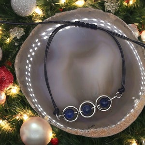 May include: A black cord necklace with three silver circles, each containing a dark blue gemstone. The necklace is displayed on a gray and white agate slice.