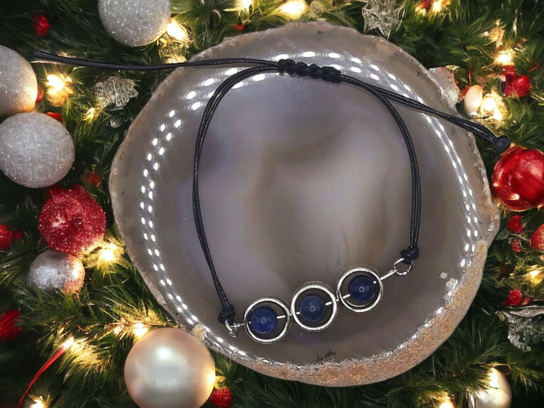 May include: A black cord necklace with three silver circles, each containing a dark blue gemstone. The necklace is displayed on a gray and white agate slice.