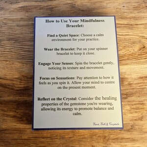 May include: A white card with blue text explaining how to use a mindfulness bracelet. The card includes instructions on finding a quiet space, wearing the bracelet, engaging your senses, focusing on sensations, and reflecting on the crystal. The card also includes the text "Taret. Talk & Crystals".