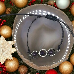 May include: A black cord bracelet with three silver rings and amethyst stones. The bracelet is displayed on a grey agate slice.