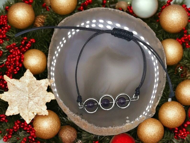 May include: A black cord bracelet with three silver rings and amethyst stones. The bracelet is displayed on a grey agate slice.